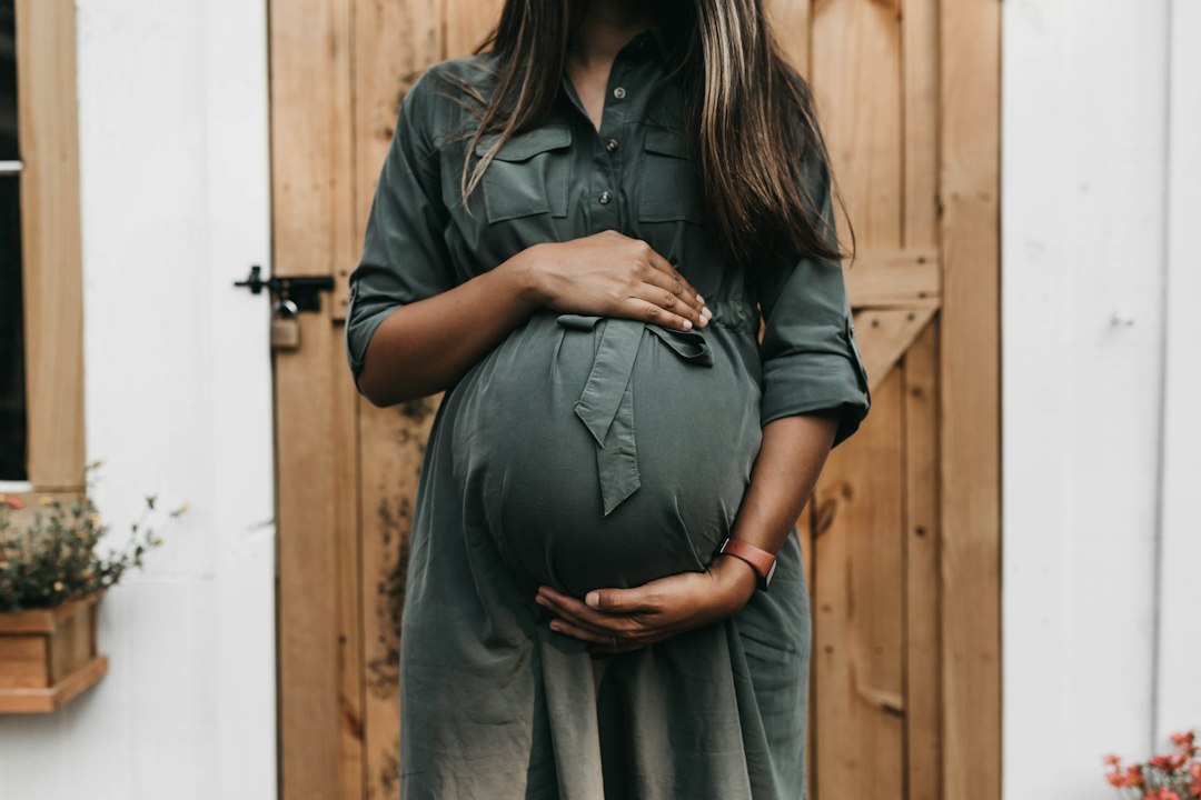 Femme enceinte en robe verte, les deux mains posées sur son ventre, devant une porte en bois